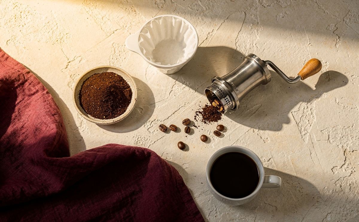 Ground coffee, a hand grinder, a pour-over cone and a cup of black coffee laid out on a pale stone counter